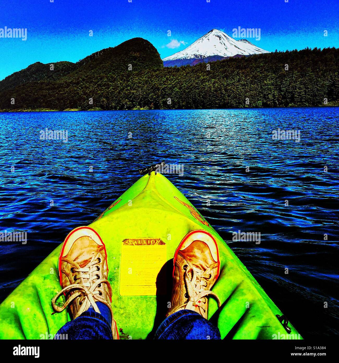 A canoeist's feet in the front section of a lime-green canoe in the foreground on a rippling lake, with a snow-capped volcano and native forest in the background. Stock Photo