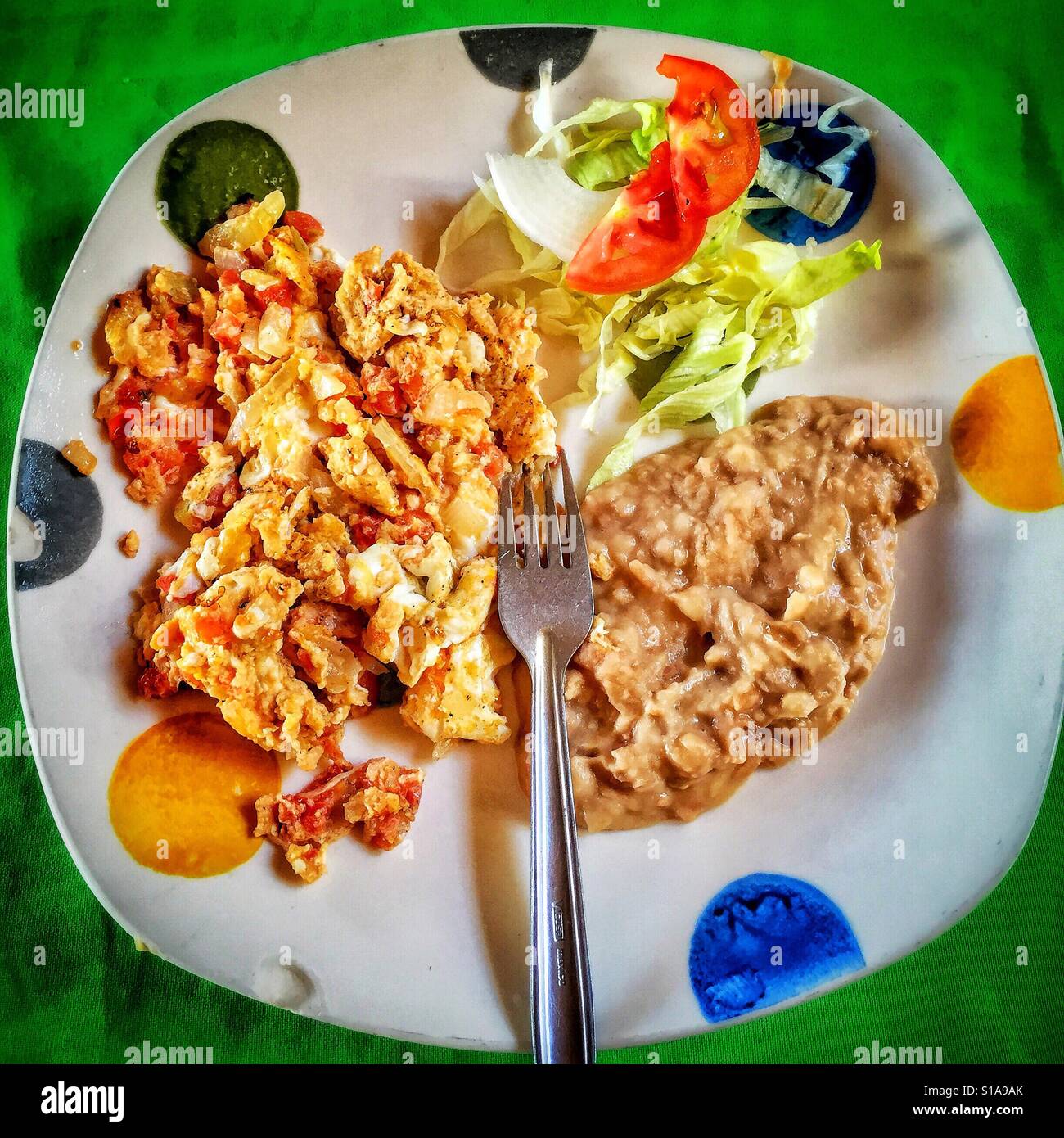 A colorful plate painted with giant polka dots is filled with a traditional Mexican breakfast of Huevos Mexicanos, refried beans, and a salad garnish. Stock Photo