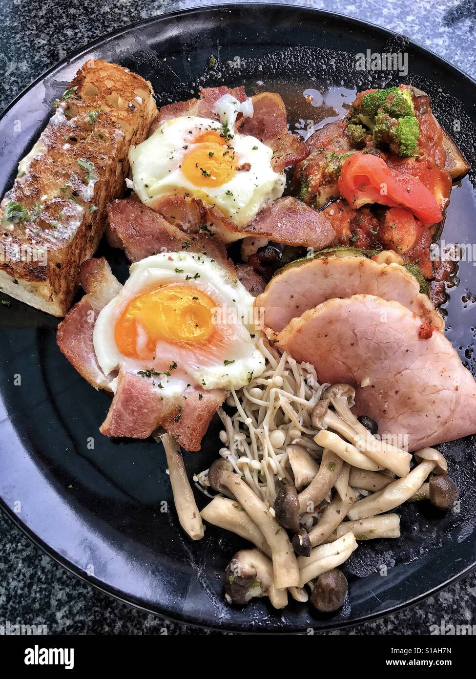 Thailand style mixed grill breakfast with eggs, bacon, ham, courgettes, mushrooms and garlic toasted bread Stock Photo
