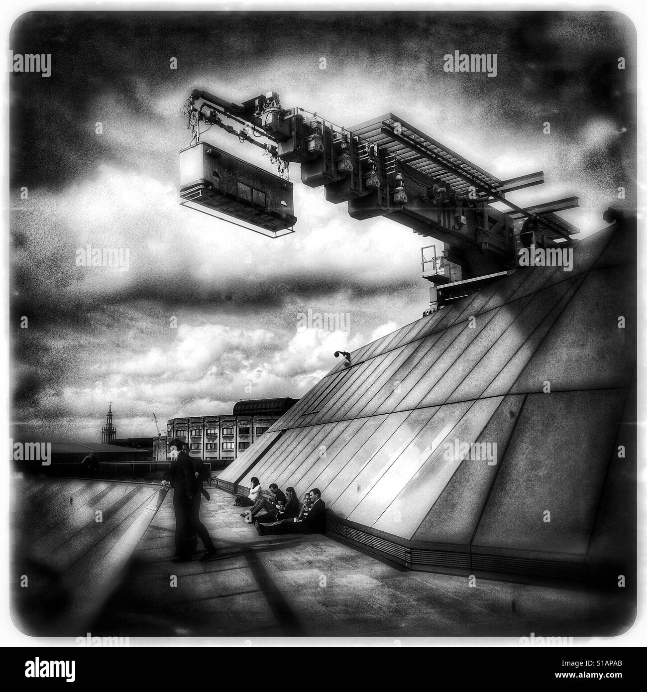 Black and white photo with a futuristic, dreamlike feel of office workers sitting as a large service crane moves overhead on the roof terrace of One New Change, London, England, UK. Stock Photo