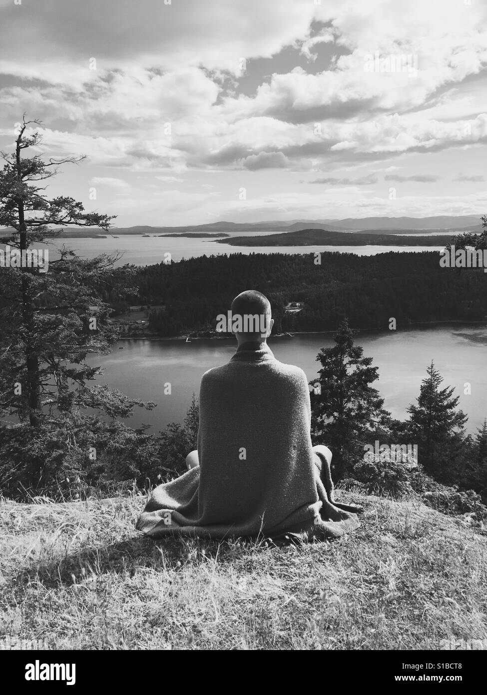 Black and white photo of person in flowing clothing with shaved head Meditating on a mountaintop overlooking the Gulf Islands Canada Stock Photo