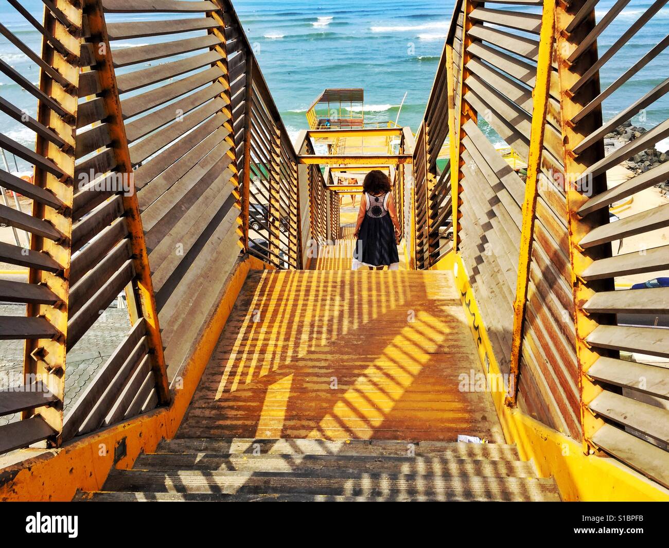 Woman in black dress walking down bright yellow stairs toward Pacific Ocean in Lima, Peru - Smartphone Captured Stock Image