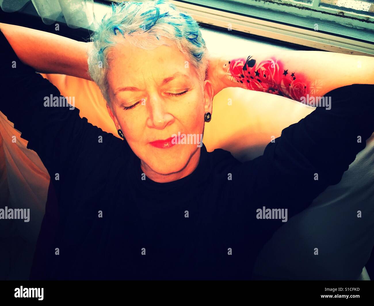 Mature woman with blue hair highlights and pink floral tattoo relaxing at home. - Smartphone Captured Stock Image