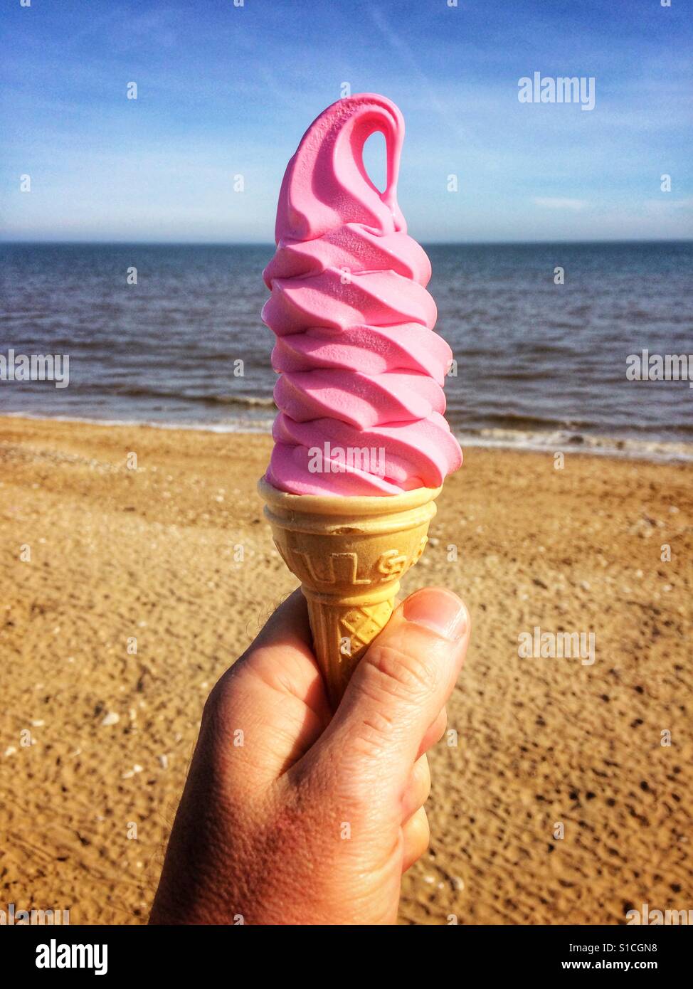 A tall Ice Cream cone with bright pink swirly ice cream being held in a hand with a beach and the sea in the background. - Smartphone Captured Stock Image