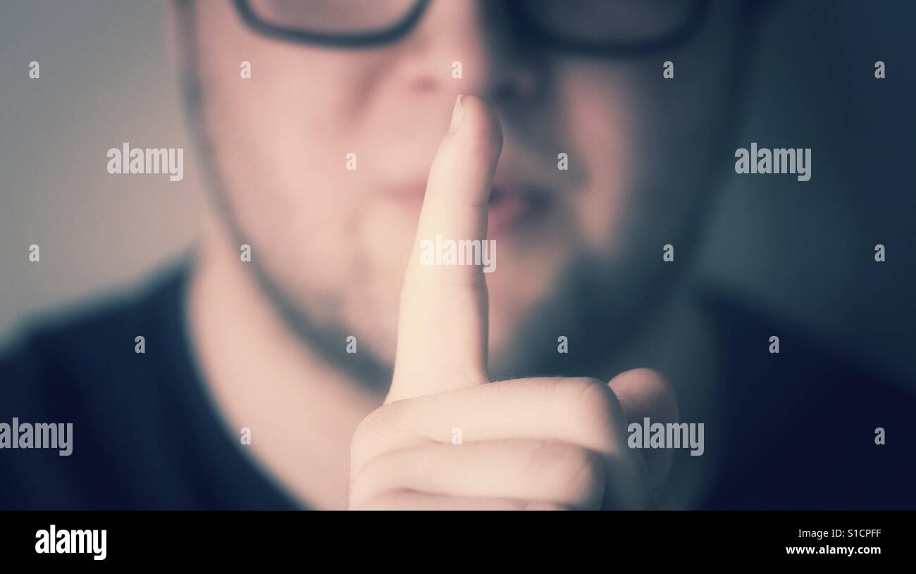 A young man holding his finger to lips in a gesture to shush or hushing another person to be quiet. Stock Photo