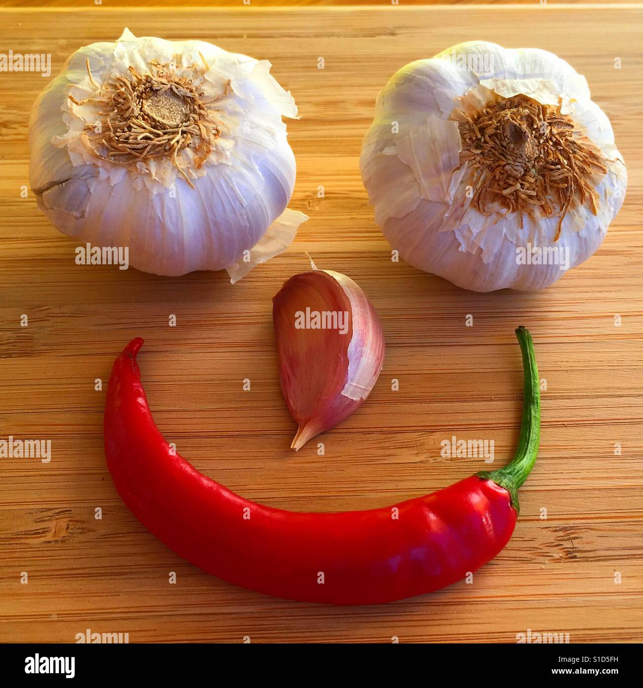 Food art. Smiley face made up from garlic bulbs, a clove  and a chilli, on a bamboo chopping board - Smartphone Captured Stock Image