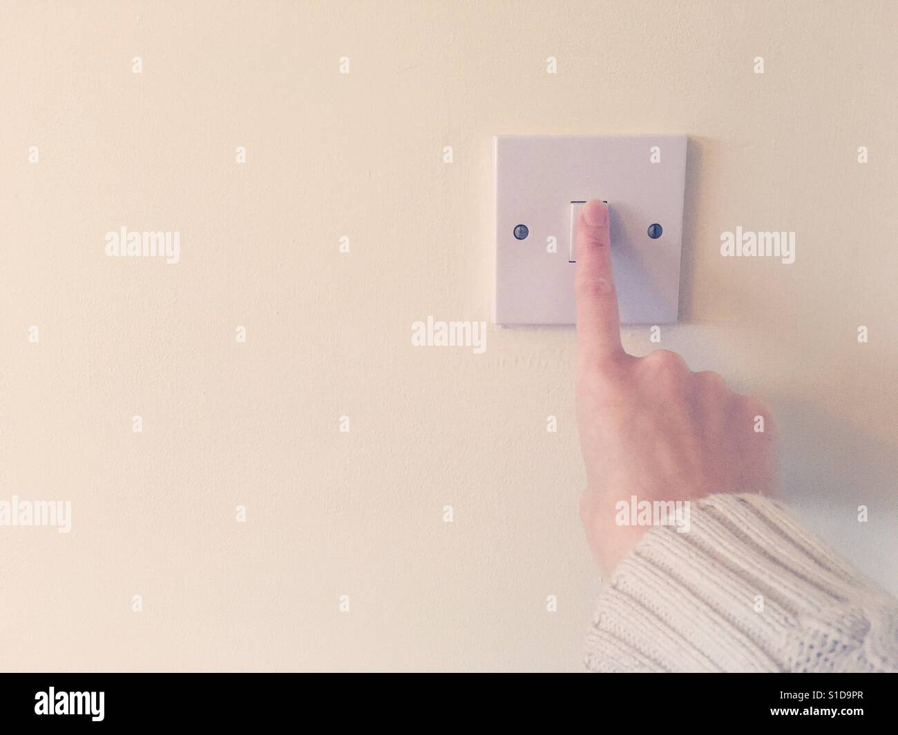 Turning on an electric light switch using index finger on right hand. Point of view perspective. Beige sleeve against magnolia coloured wall. Stock Photo