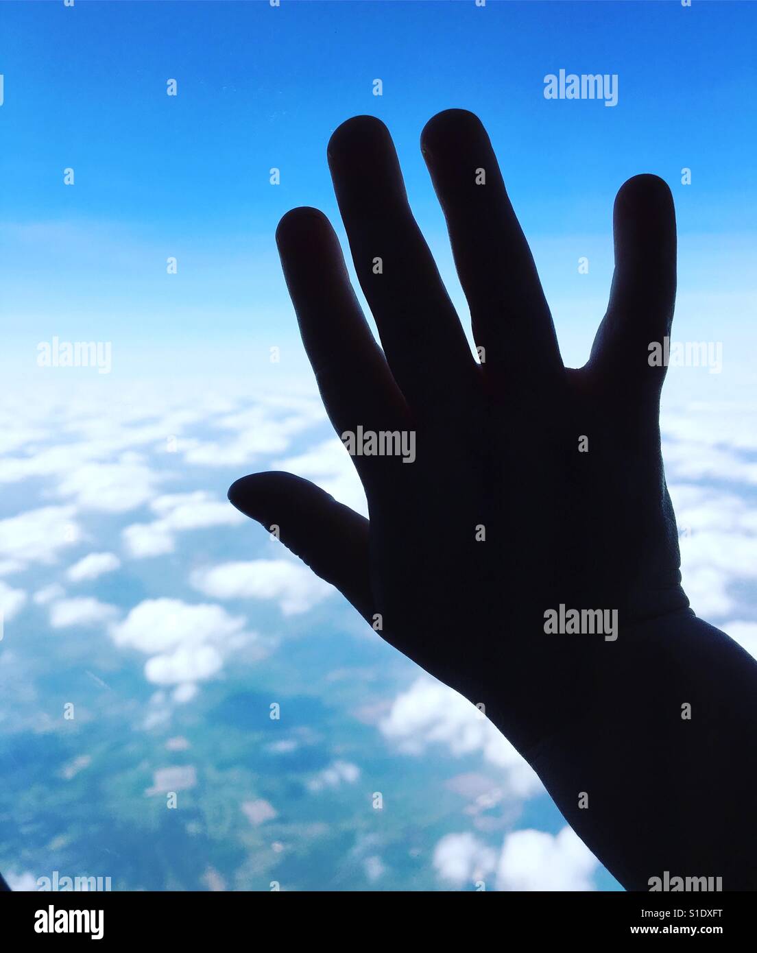 A child's hand up against the window of an airplane, silhouetted against a blue sky with white fluffy clouds Stock Photo