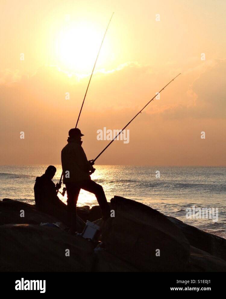 A pair of fishermen angling on the rocks of a beautiful beach at sunrise. Stock Photo
