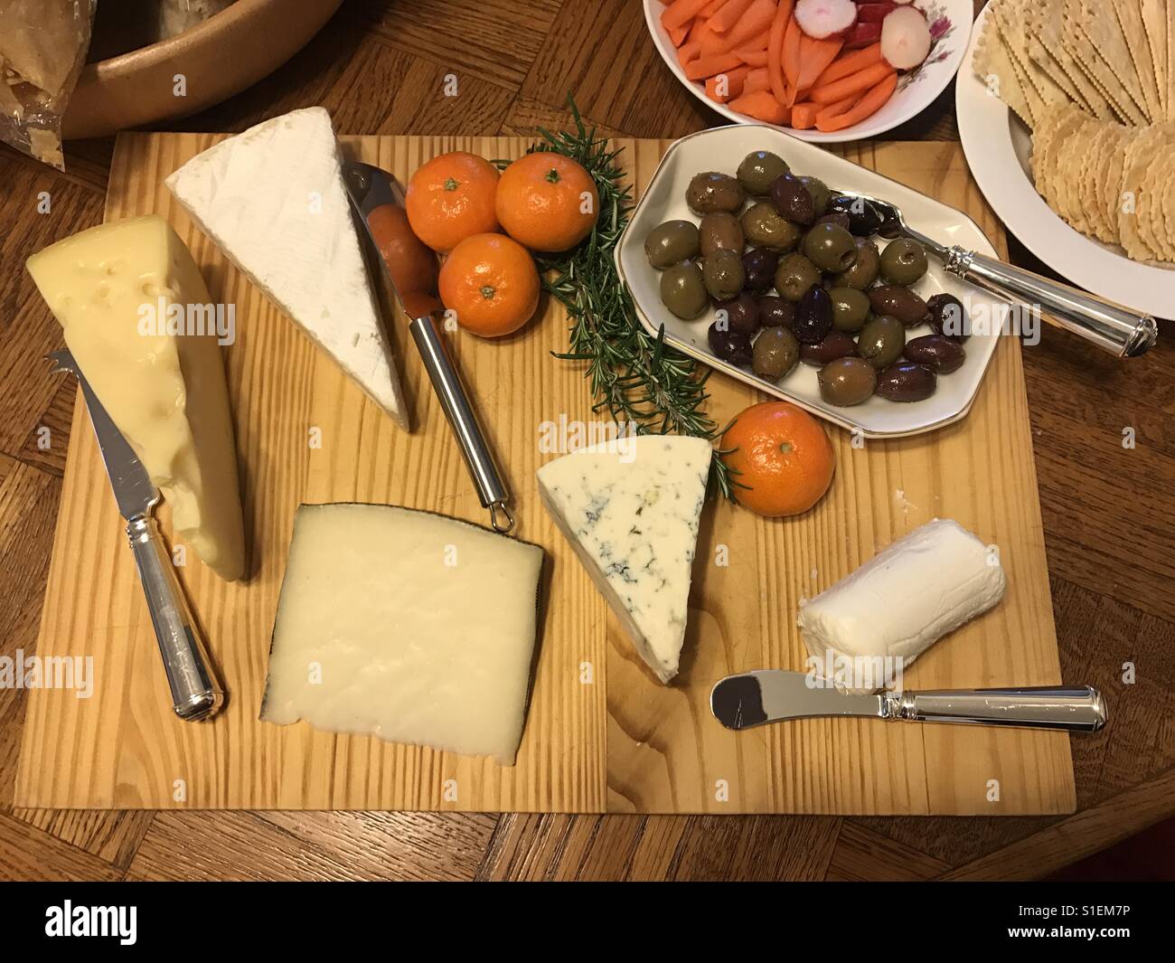 Assortment of cheeses, with dish of olives and few tangerines on wooden serving board with knives and sprig of rosemary. From above perspective. Stock Photo