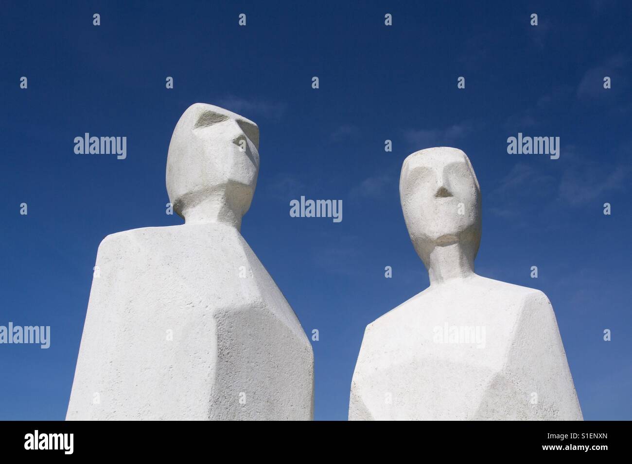 A pair of white, faceless statues including a man and a woman looking upwards to a blue sky. - Smartphone Captured Stock Image