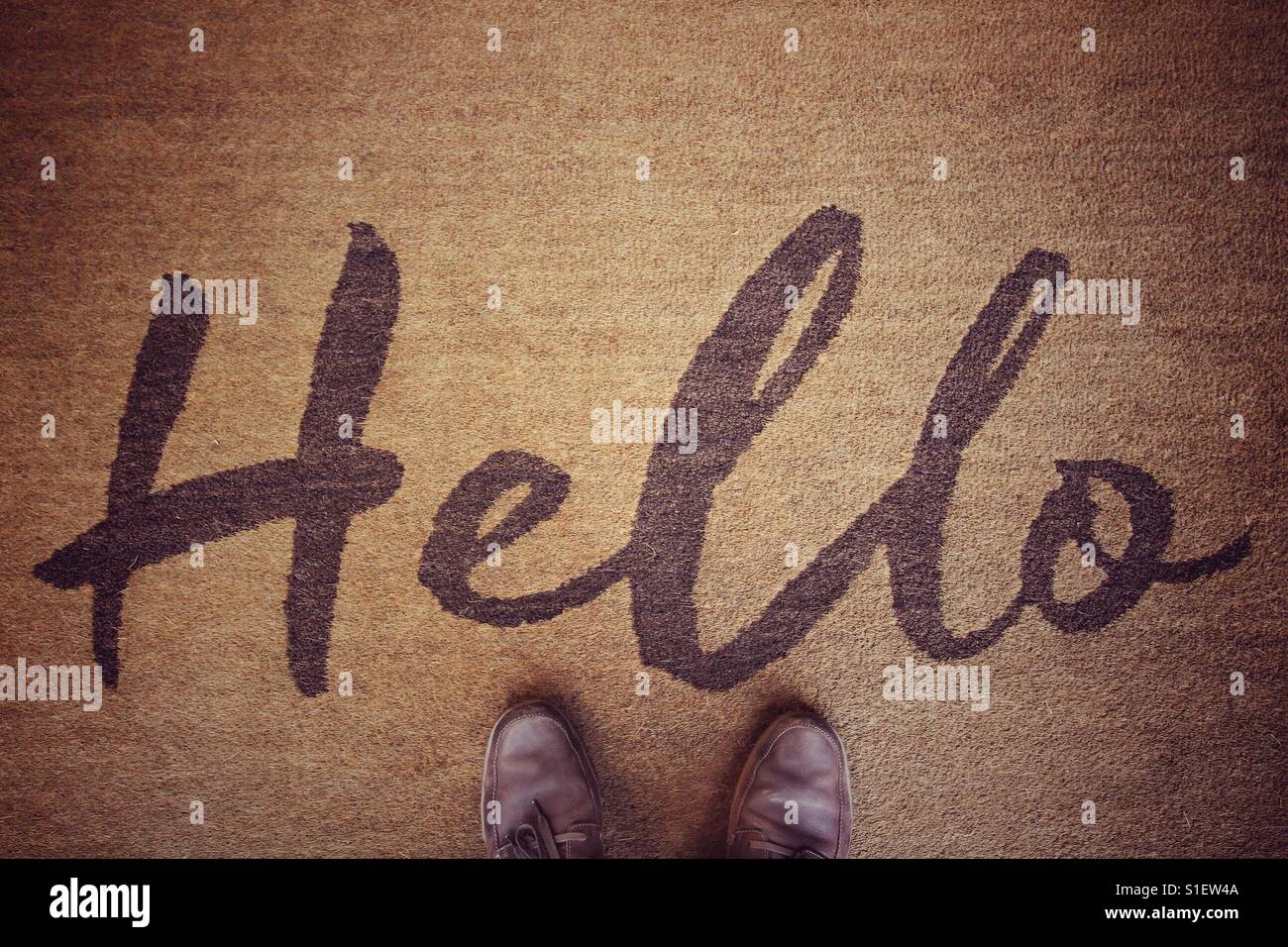 Looking down from above onto a pair of shoes that are stood on a welcome mat with the word hello written in elaborate script. Stock Photo