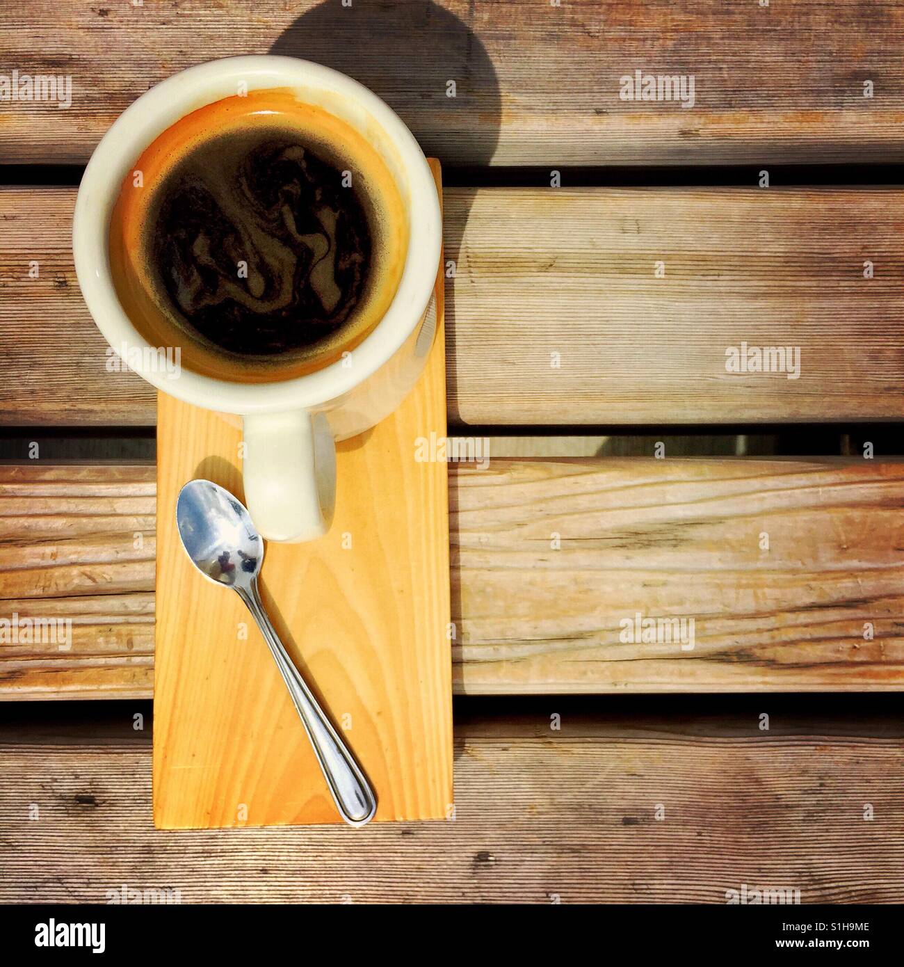 An  overhead shot of an Americano and a spoon on a wood serving plate on a wooden bench Stock Photo