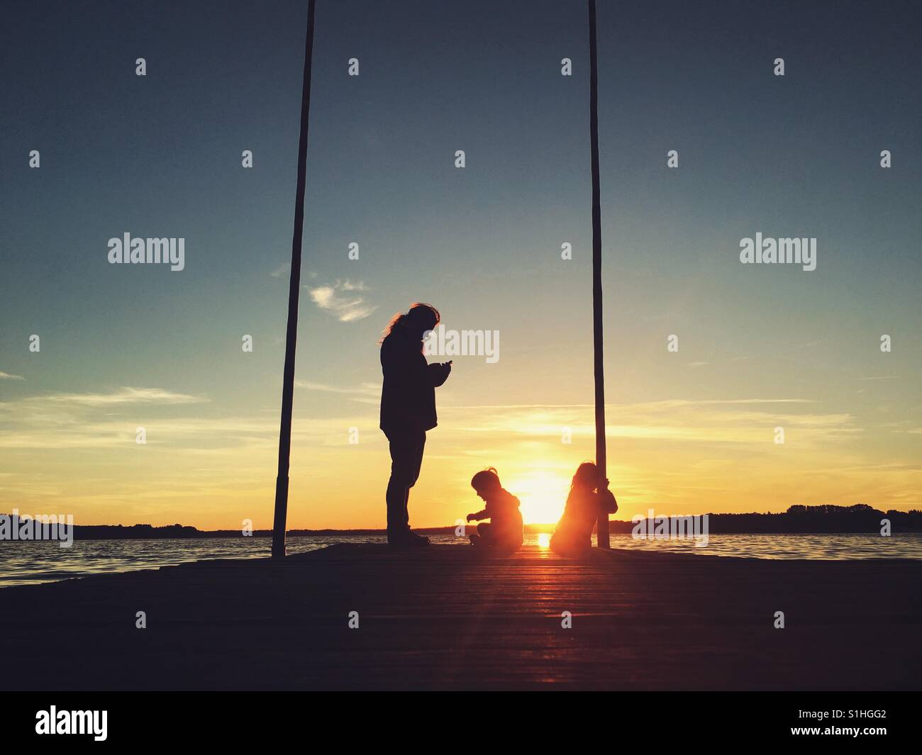 A man and two children on a jetty, the man is looking at his smartphone - Smartphone Captured Stock Image