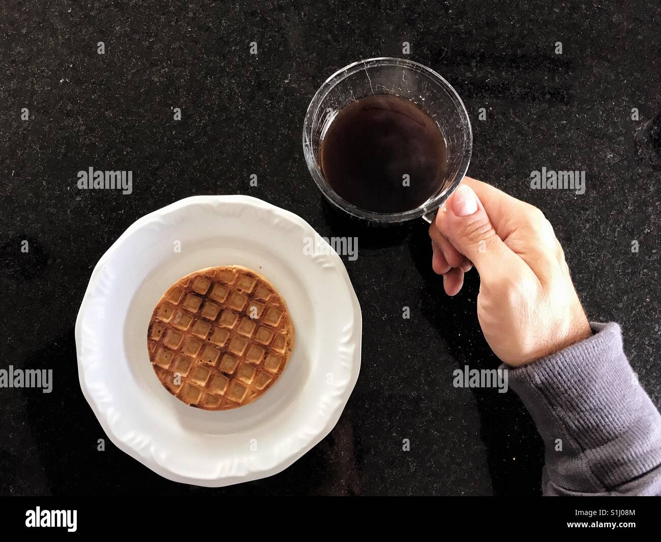 Hand holding a coffee cup and a white plate with a waffle on the granite countertop - Smartphone Captured Stock Image