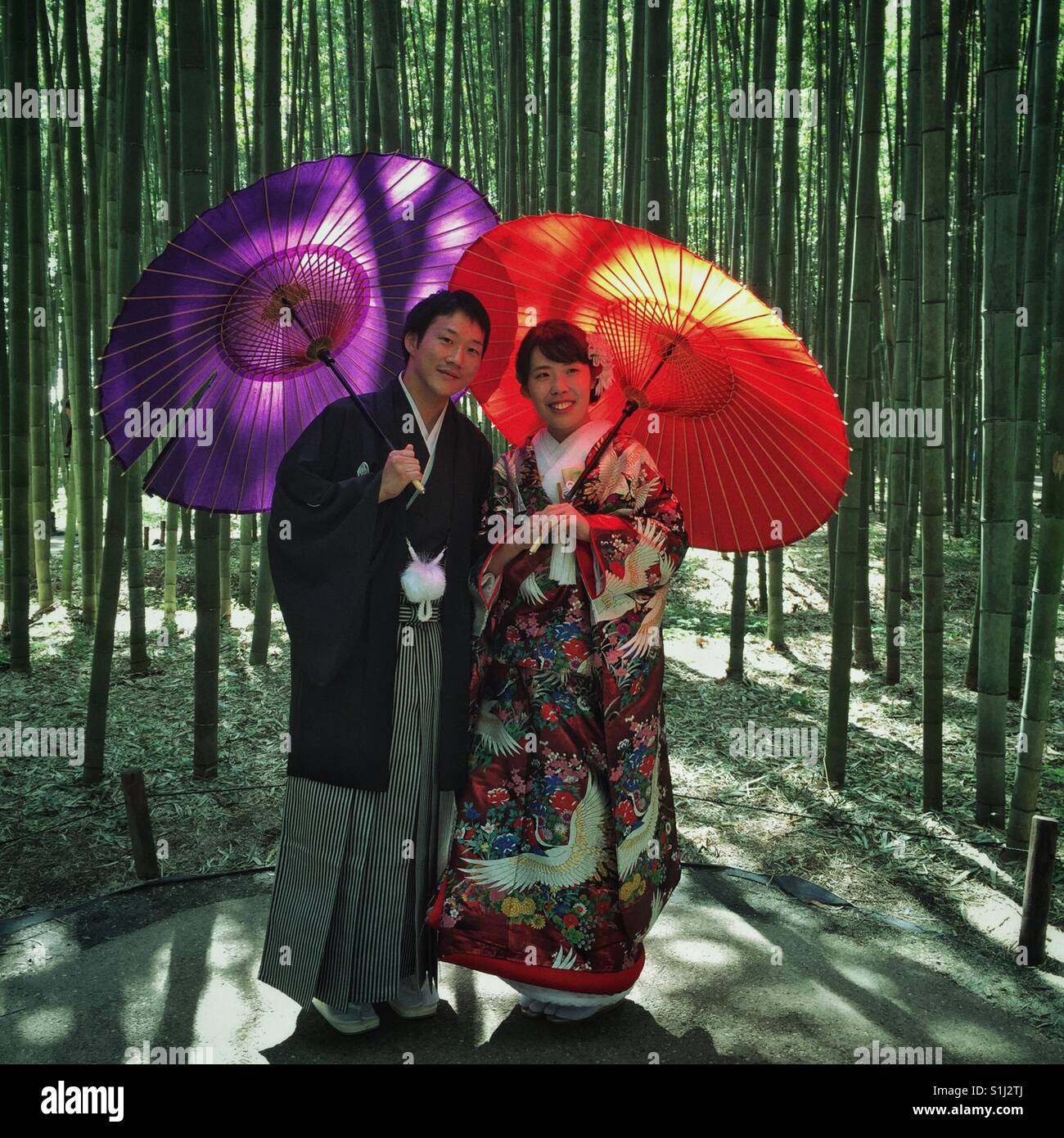 Traditionally dressed Japanese couple with red and purple umbrellas posing in front of bamboo forest in Arashiyama,Kyoto Stock Photo