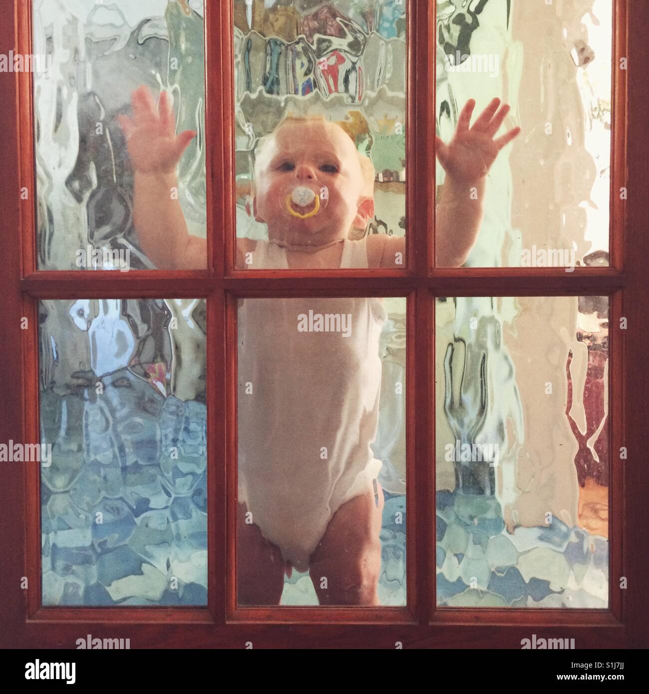Ten month old baby boy standing against a glass internal door. - Smartphone Captured Stock Image