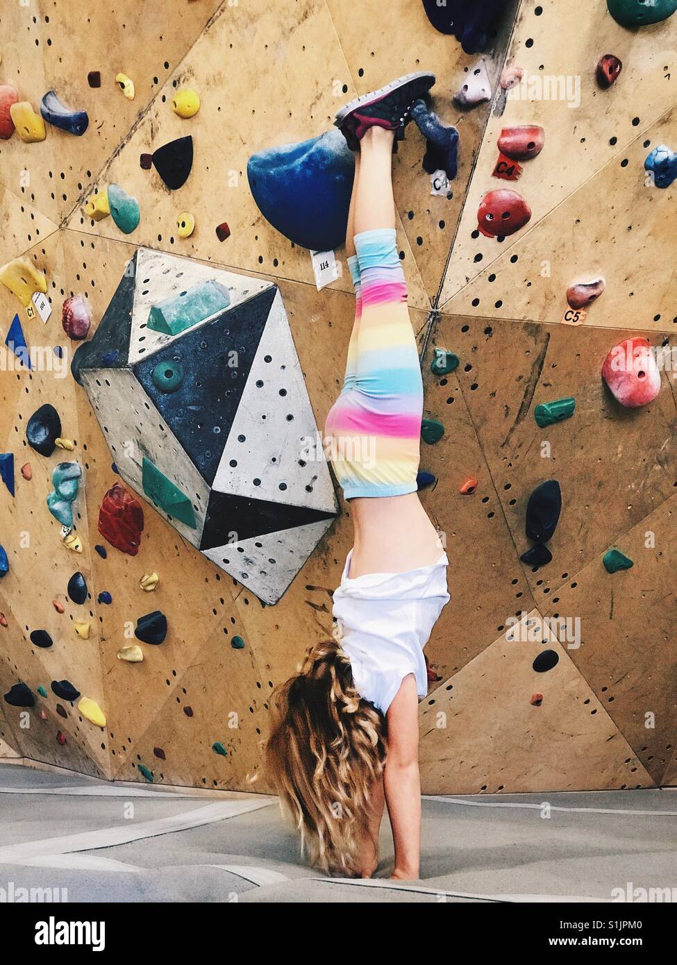 A girl does a handstand in front of an indoor bouldering wall. Stock Photo