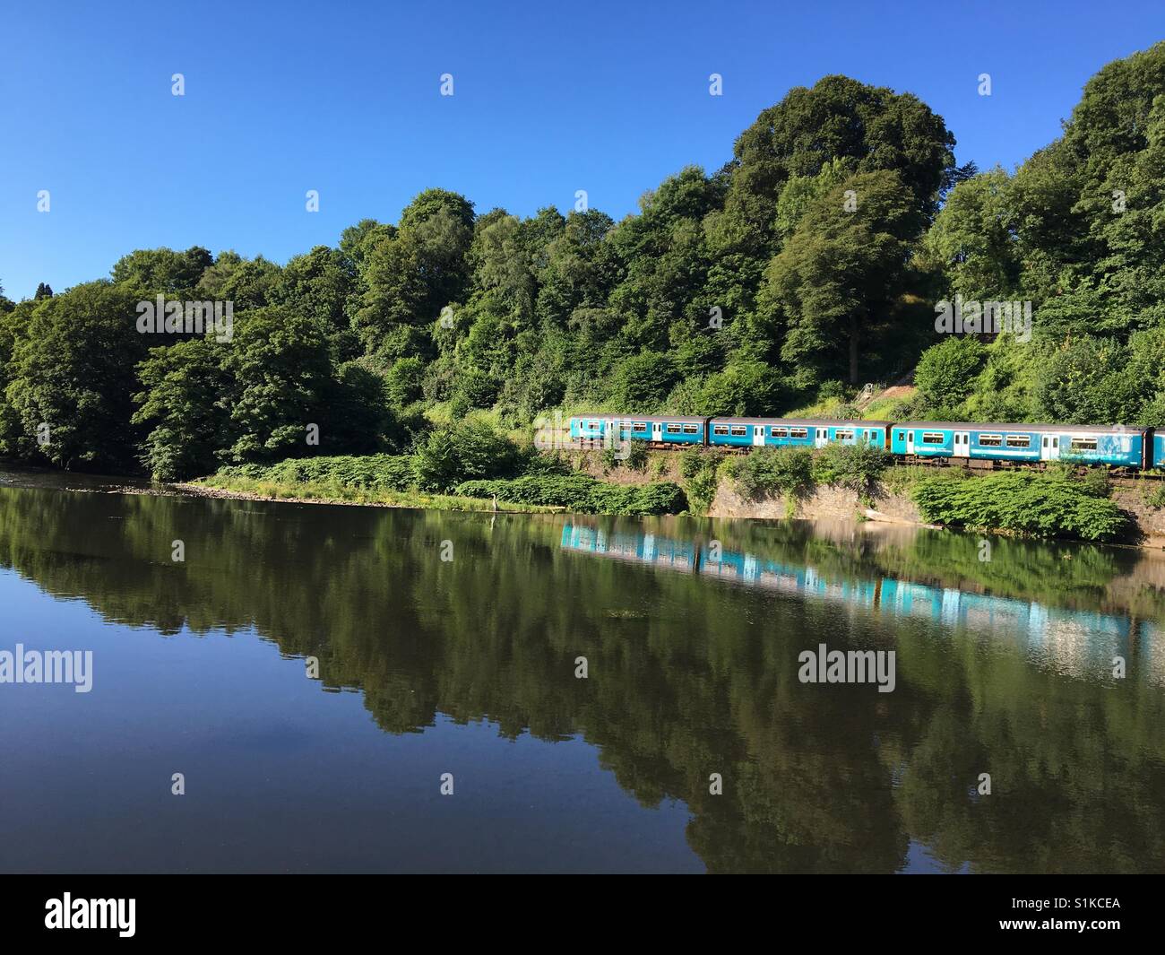 Commuter train on its journey to Cardiff early on a bright summer morning passes the still water above the weir in the River Taff at Radyr Stock Photo