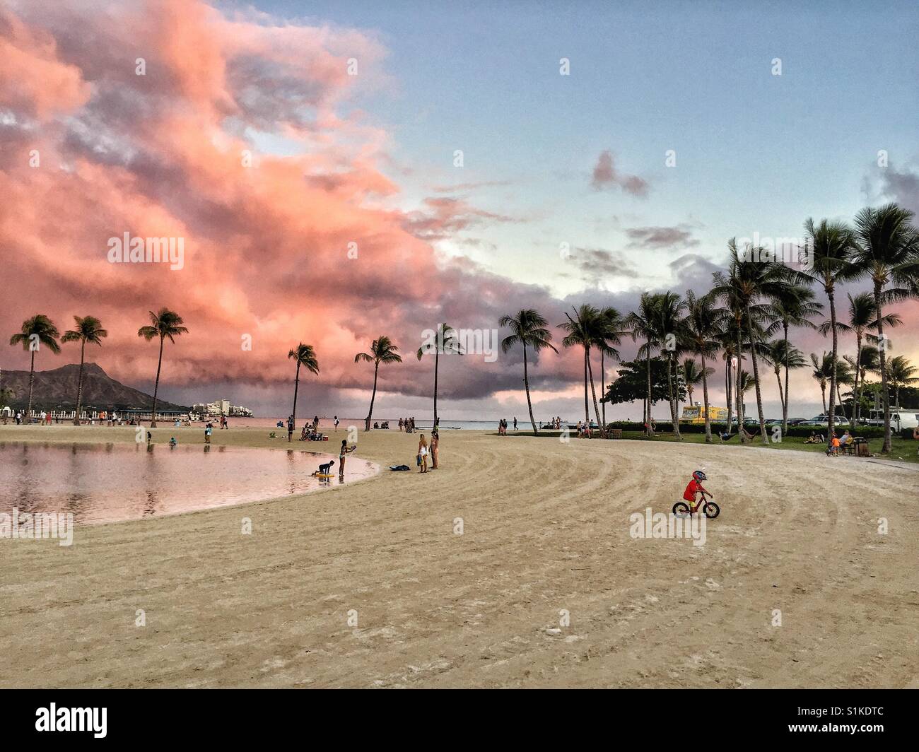 Waikiki beach and Diamond Head with pinks storm clouds overhead provide beautiful backdrop to small boy riding bicycle on sand by tropic palm tree lined lagoon, Oahu, Hawaii Stock Photo