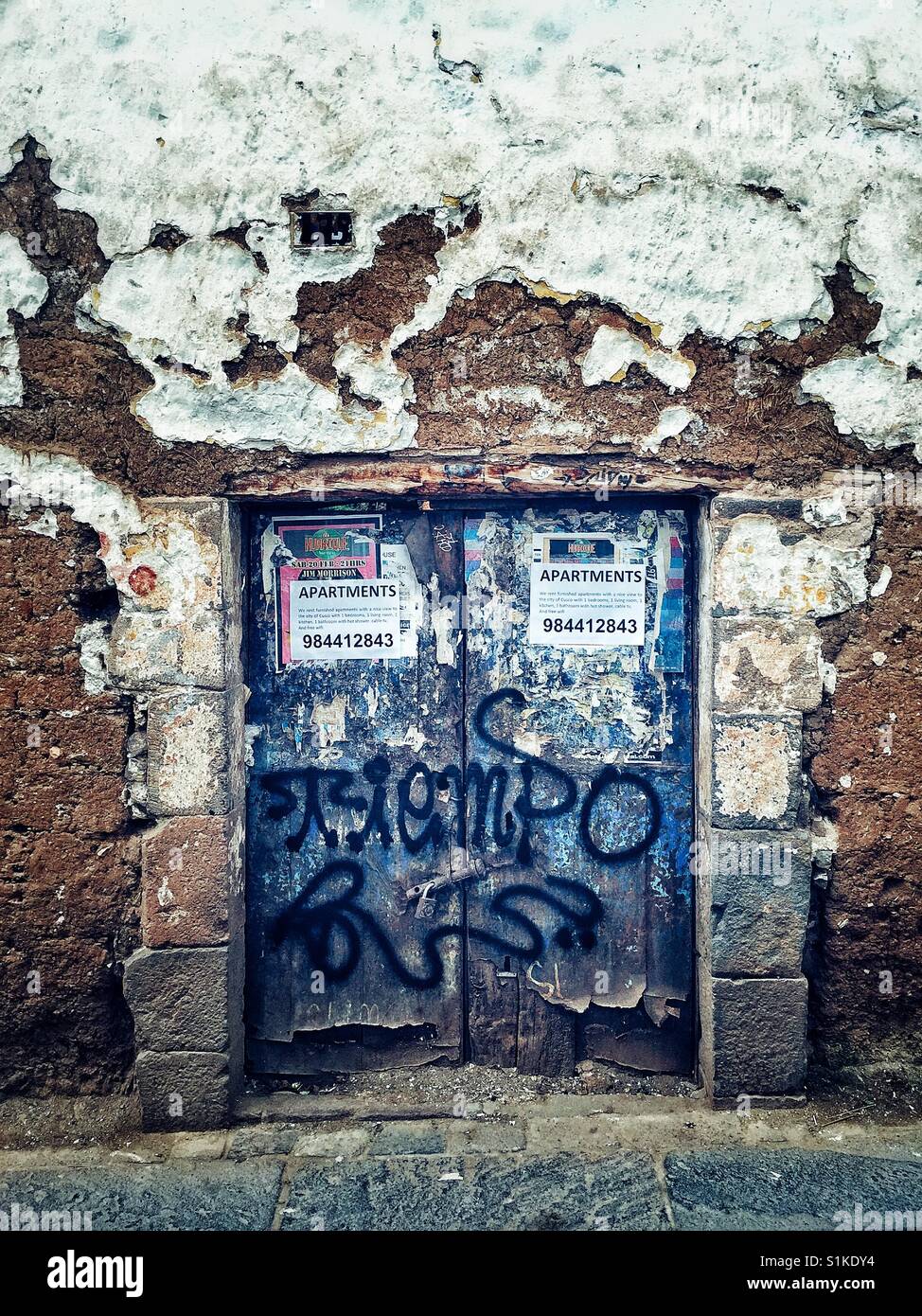 Old blue door spray painted with graffiti and plastered with ads in the scruffy  white stucco wall on the street of cusco, Peru Stock Photo