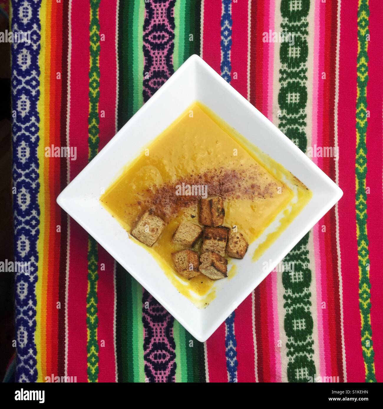 Bright yellow butternut squash soup in square dish with croutons on traditional Peruvian woven rug - Smartphone Captured Stock Image