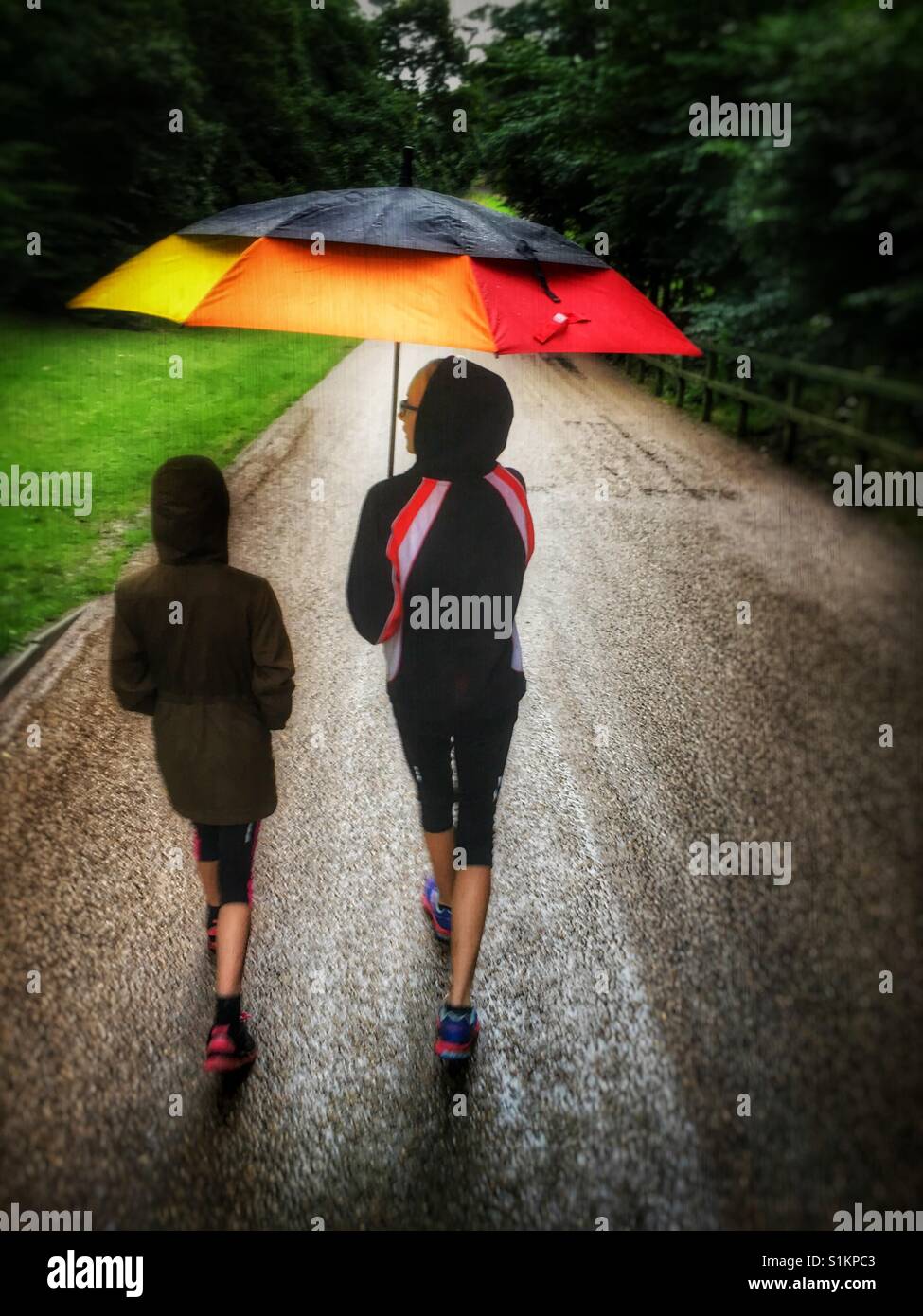 Two young girls walking in rain under a large colourful umbrella Stock