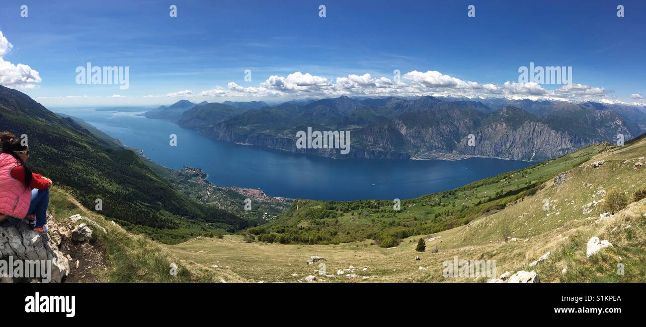 Lake Garda, Italy - May 2017: Panoramic view from Monte Baldo looking down to Malcesine with a visitor sitting on a rock - Smartphone Captured Stock Image