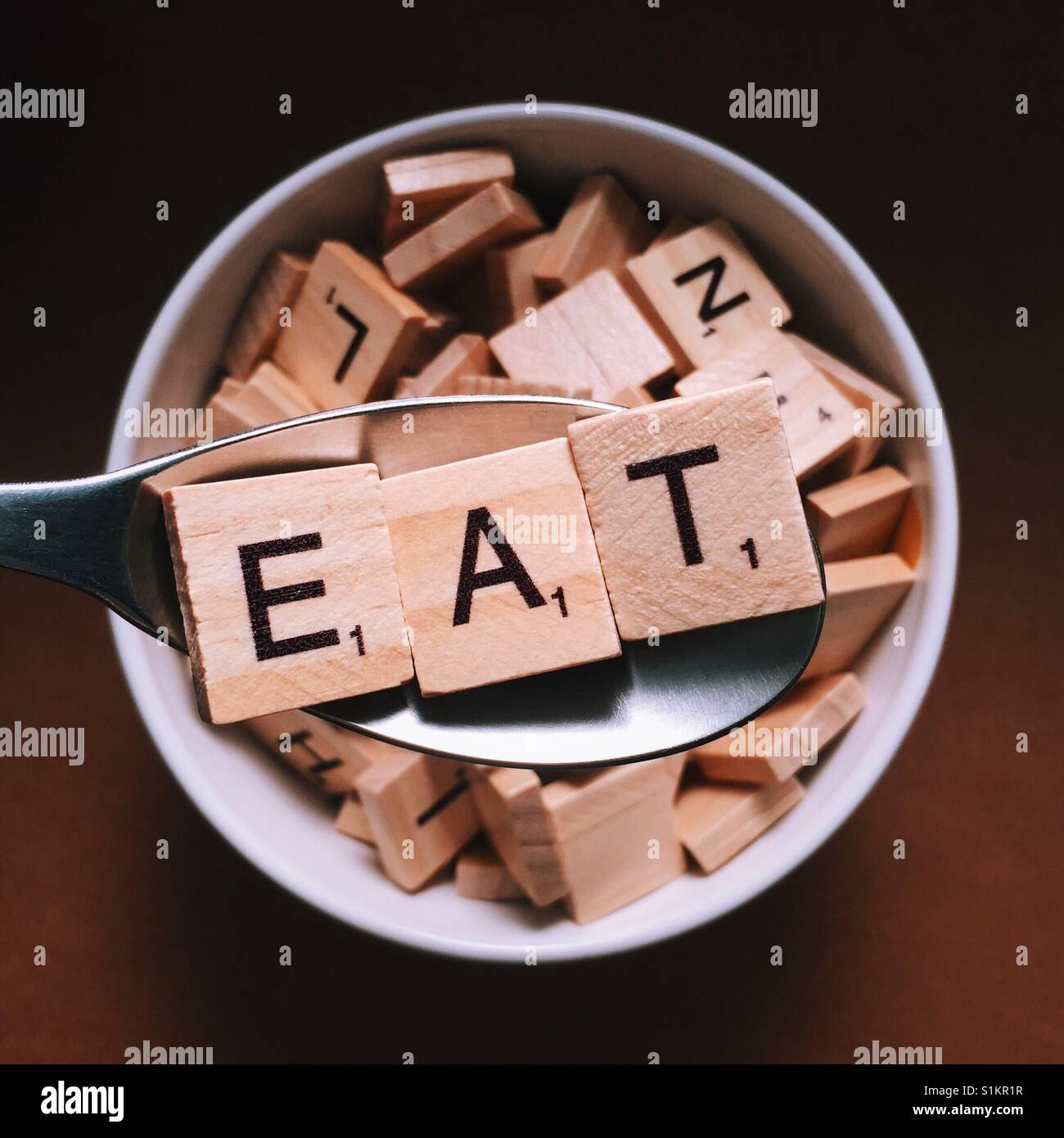 Close up of a spoon with wooden letters on it spelling eat and a bowl full of wooden letters beneath it Stock Photo