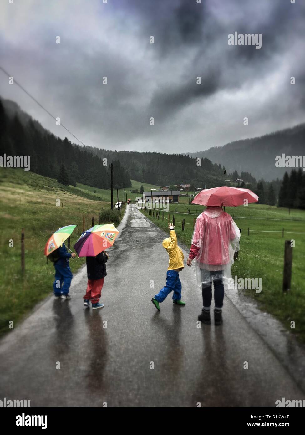 Kids and a woman in raingear walking down a Street in the countryside - Smartphone Captured Stock Image