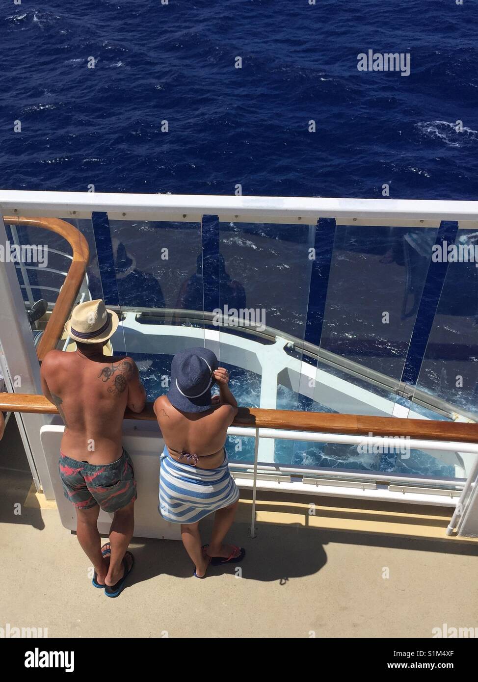 Couple in 20-30s on cruise ship deck wearing swim suits and hats enjoying the view as ship departs port. Looking down In man and woman lean on deck railing of cruise line. Tropical voyage Stock Photo