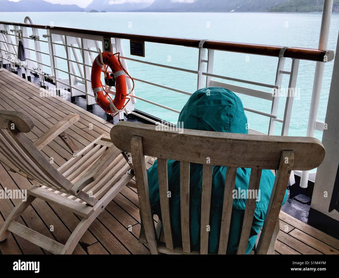 Person seats on wooden deck chair on outside deck of cruise ship in rain jacket enjoying scenic view of coastline on cold day. Glacier Bay, Alaska Stock Photo