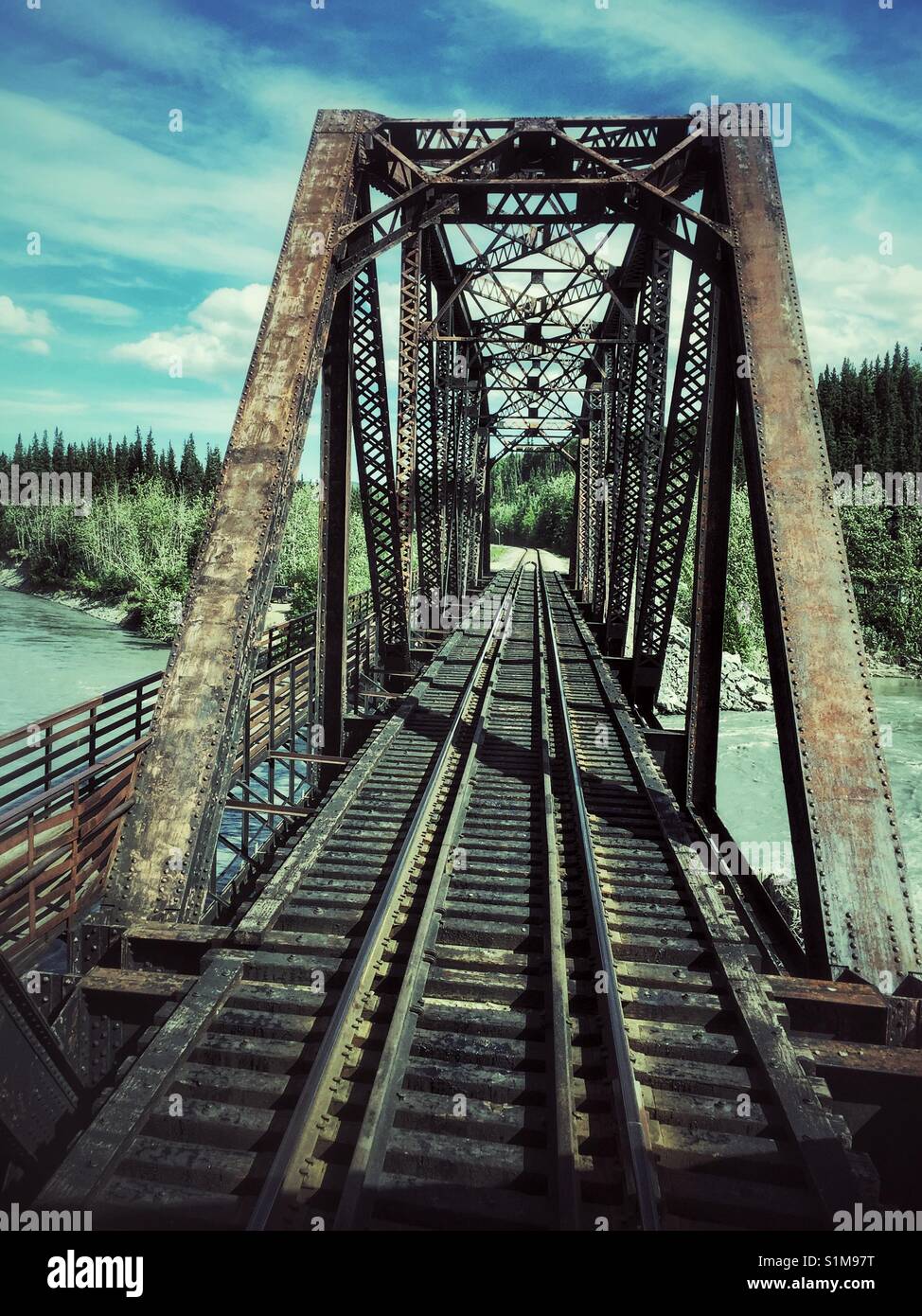 Train tracks leading across metal railway bridge to trees on distance shore. Steel railway bridge between Fairbanks and Denali, Alaska Stock Photo