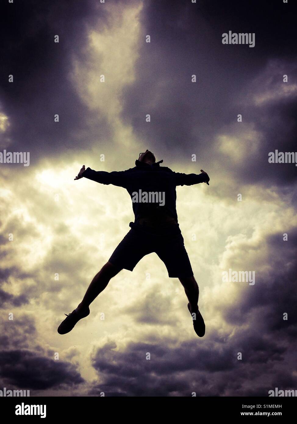 A man jumping high in the air with his arms and legs spread as if falling with a dramatic, stormy sky behind Stock Photo
