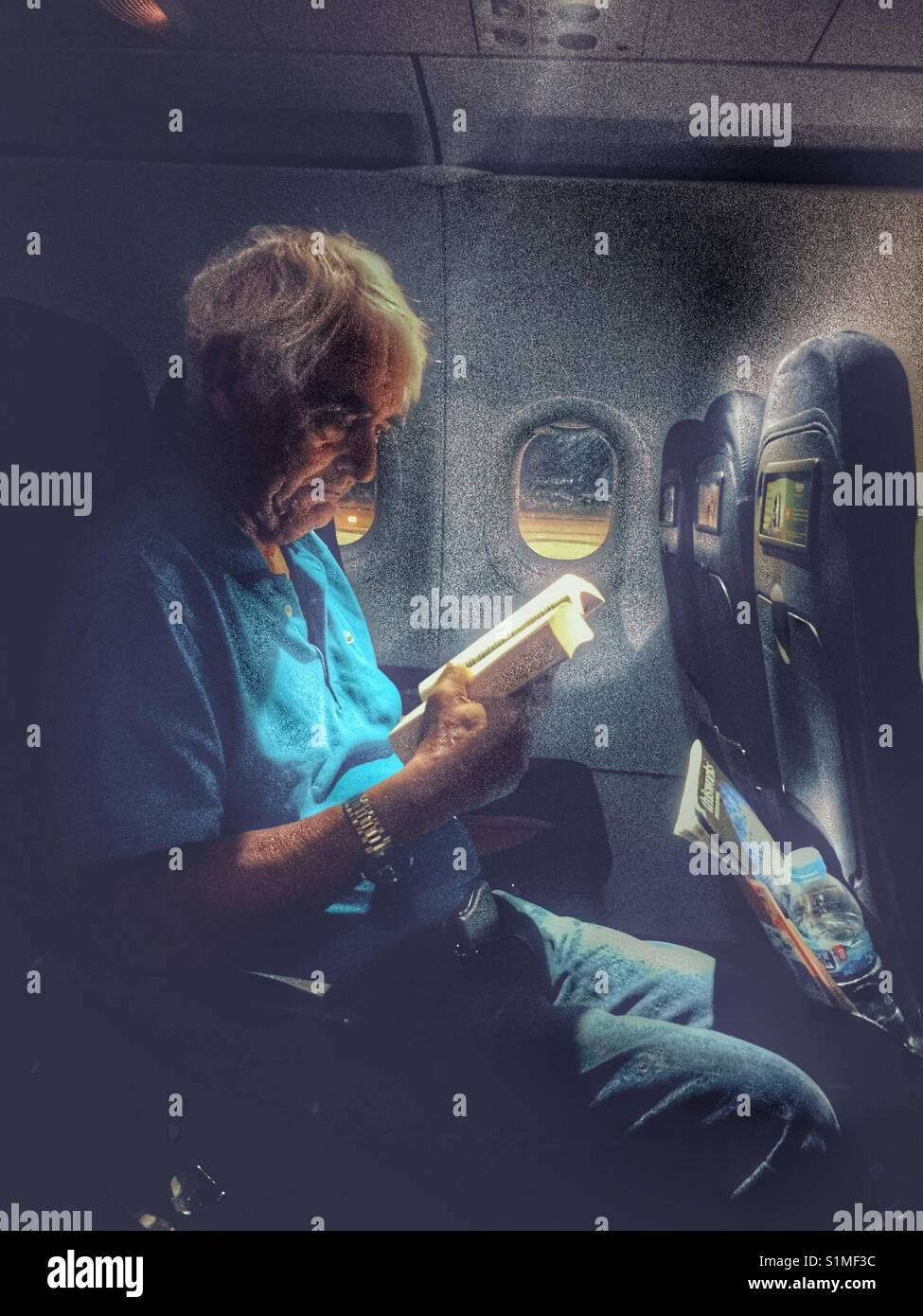 Senior man reading a book on an aeroplane at night, waiting for take-off - Smartphone Captured Stock Image