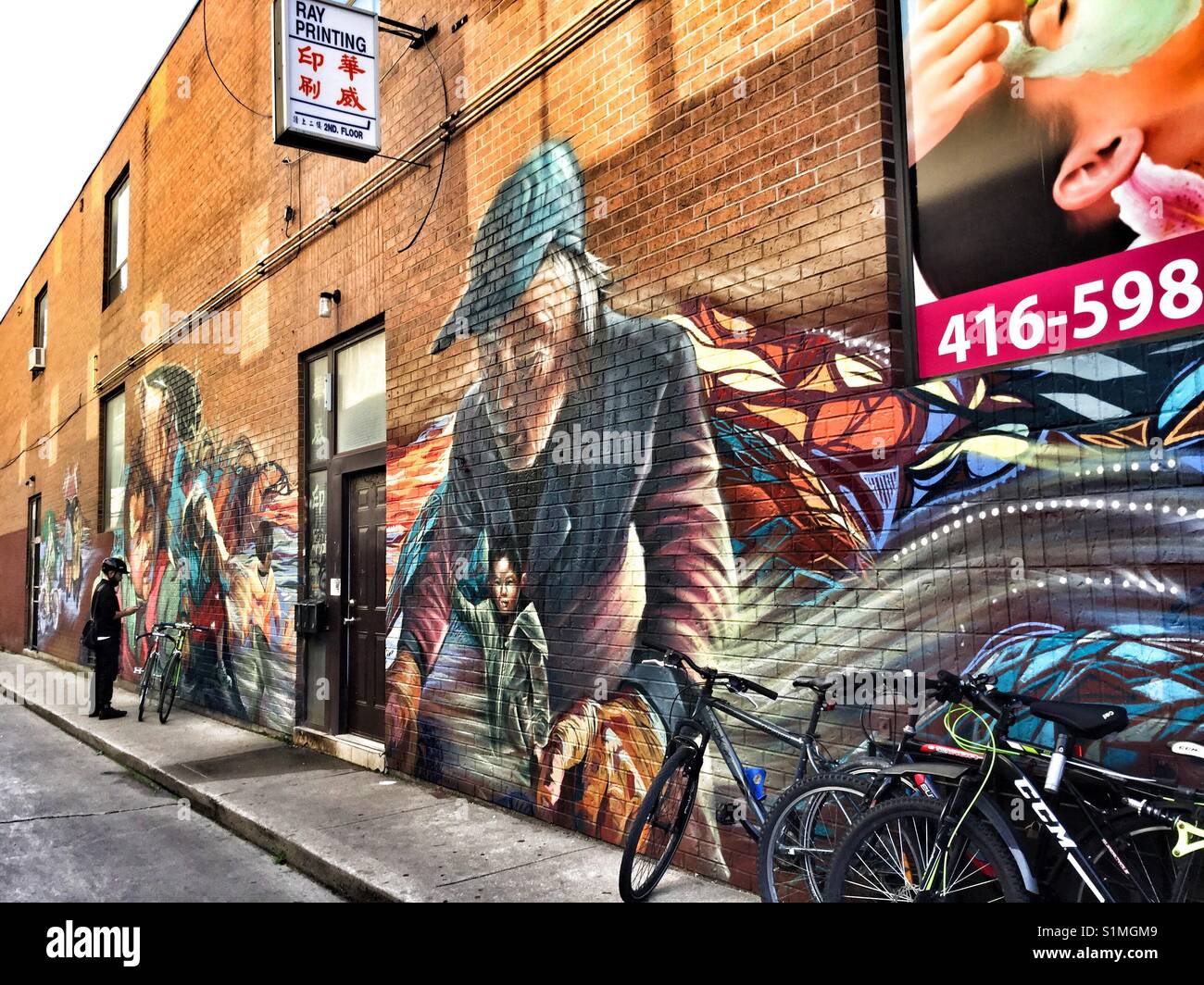 Man stands beside his bike next to a large painted brick wall. - Smartphone Captured Stock Image