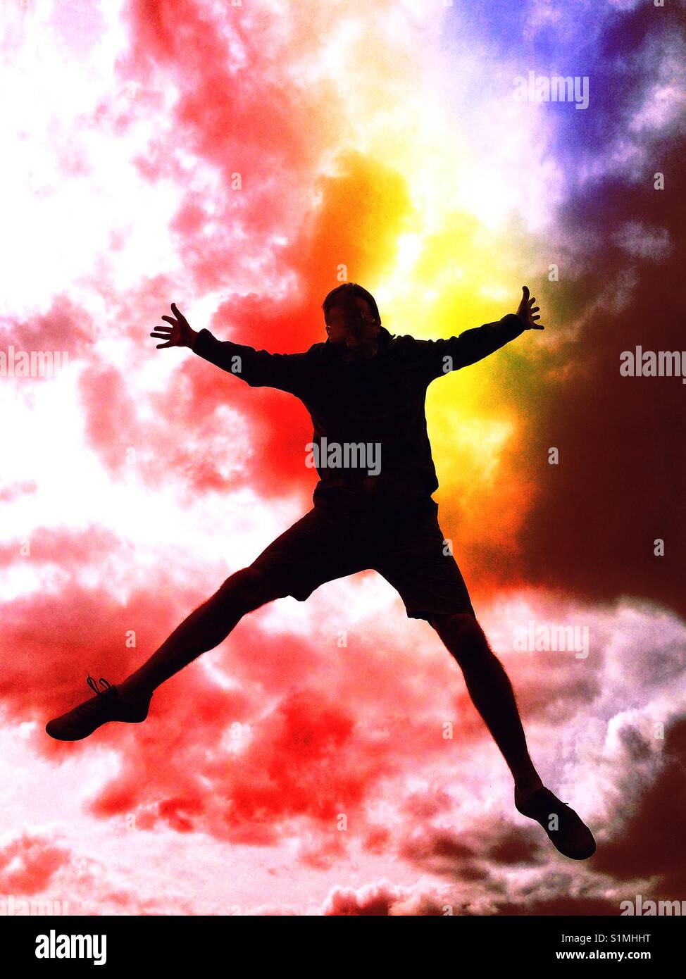 The silhouette of a man jumping high in the air with his arms and legs splayed and a colourful stormy and cloudy sky behind him - Smartphone Captured Stock Image