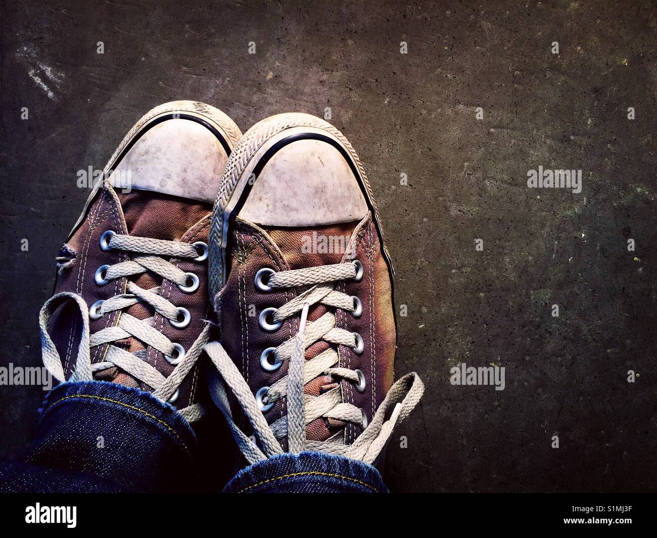 Close up of an old pair of canvas sneakers on a concrete surface being worn by a man in jeans Stock Photo
