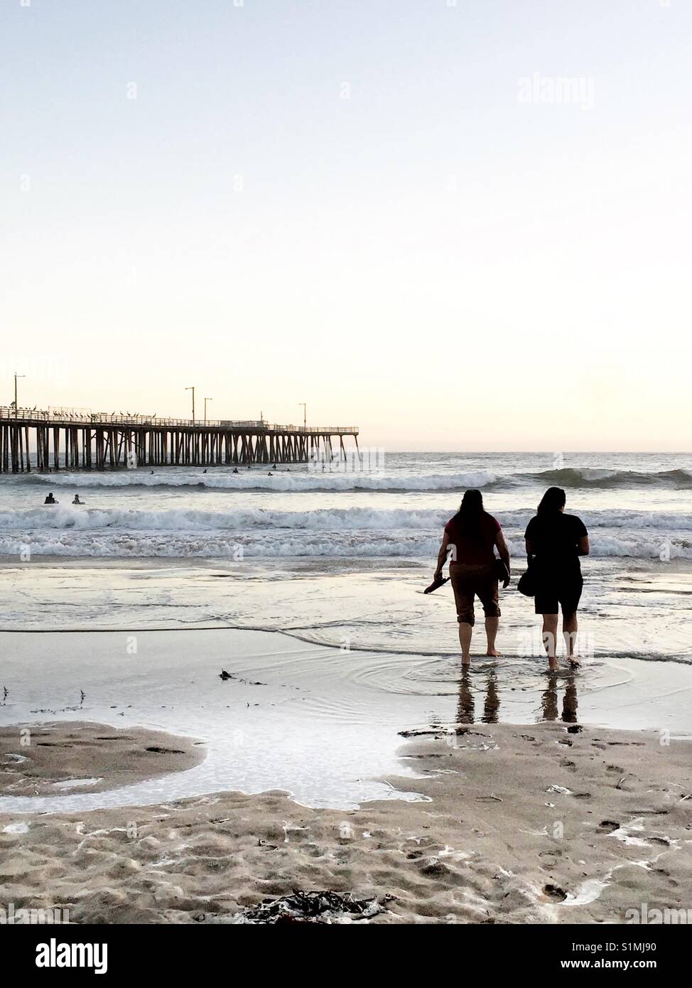 Two women step into the water during their evening beach walk. Scenic California seaside with iconic long pier and silhouette of two women at waters edge. Stock Photo