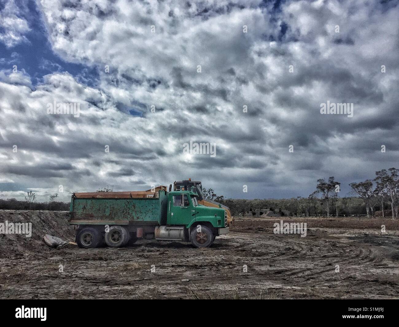 Tip trucks parked on muddy ground at cleared building site. Background of Australian bushland and storm clouds. Stock Photo