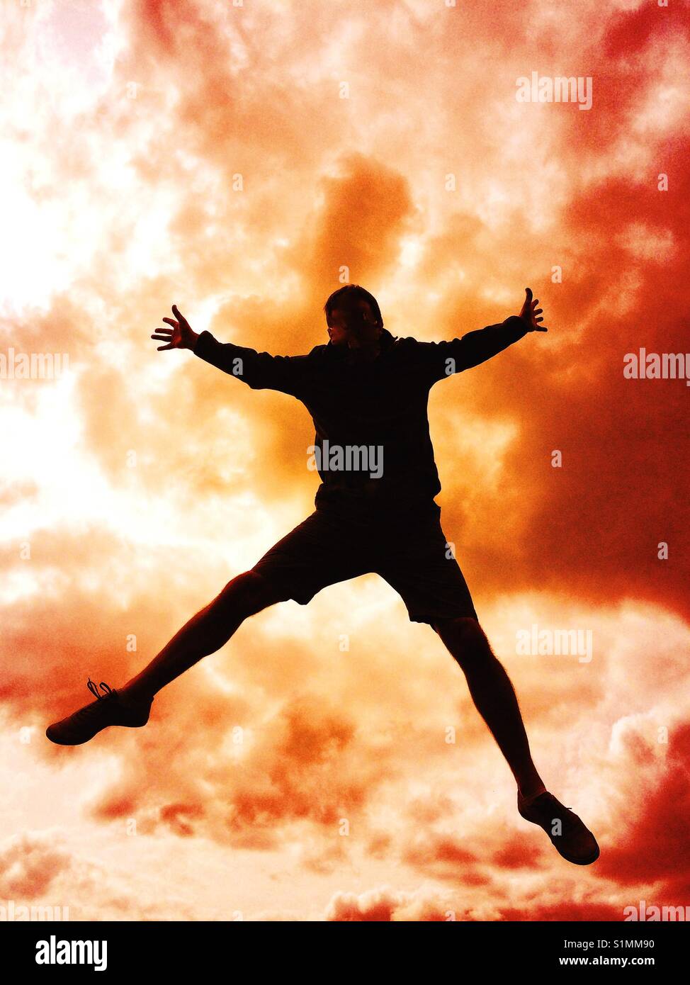 The silhouette of a man jumping high in the air of falling from the sky with a stormy sky behind that looks like an explosion or ball of flames - Smartphone Captured Stock Image