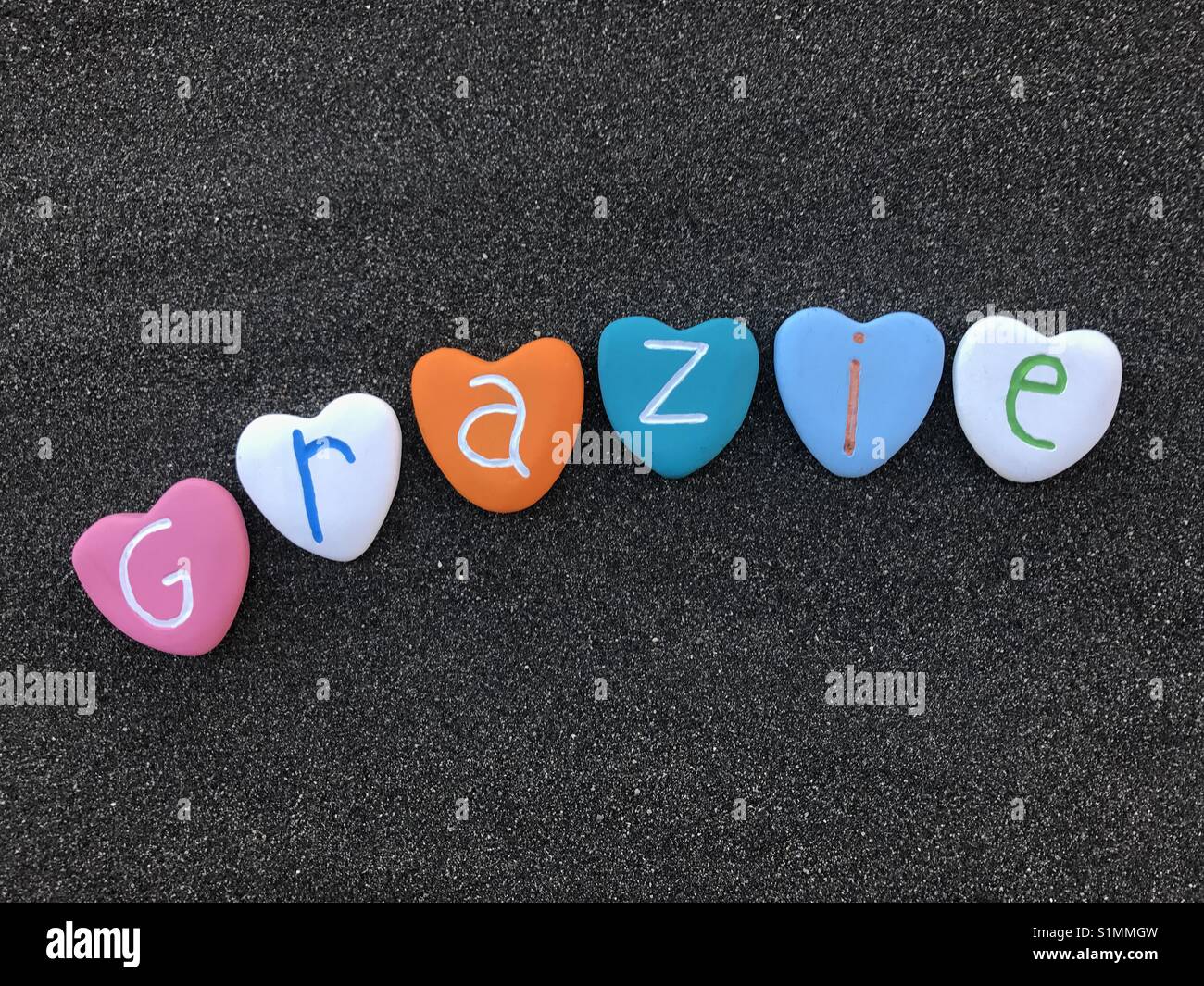 Grazie, italian thank you with colored heart stones over black volcanic sand Stock Photo