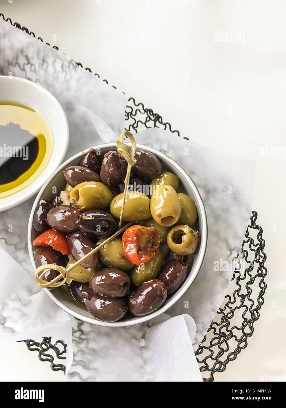 Small white dish of juicy green and black olives and sun dried tomatoes set in wire basket with olive oil and balsamic vinegar on the side. - Smartphone Captured Stock Image