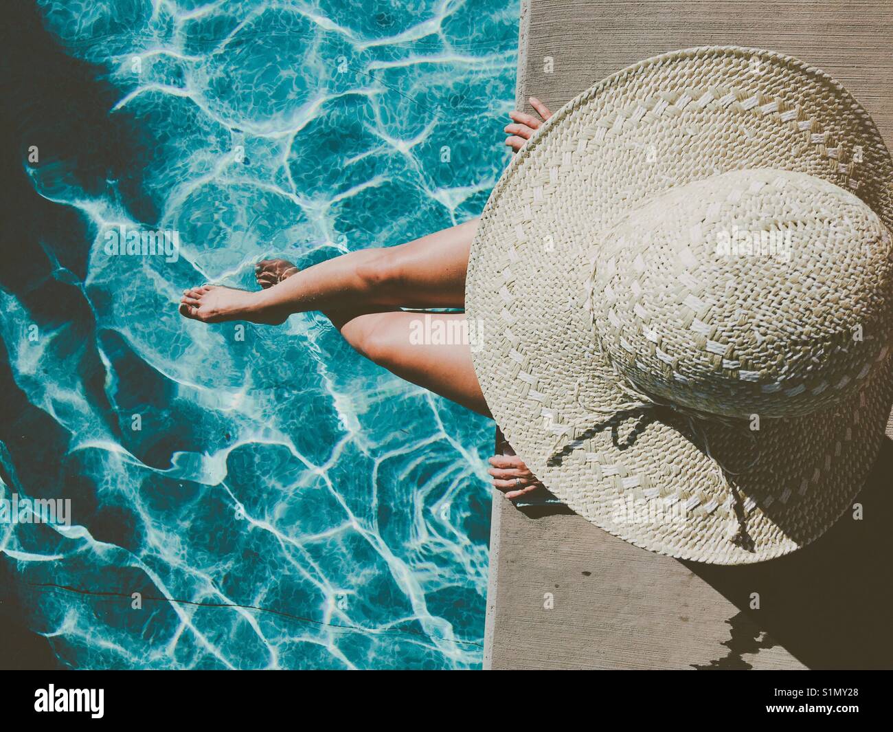 Woman wearing a large sun hat sitting at the edge of swimming pool on a ...
