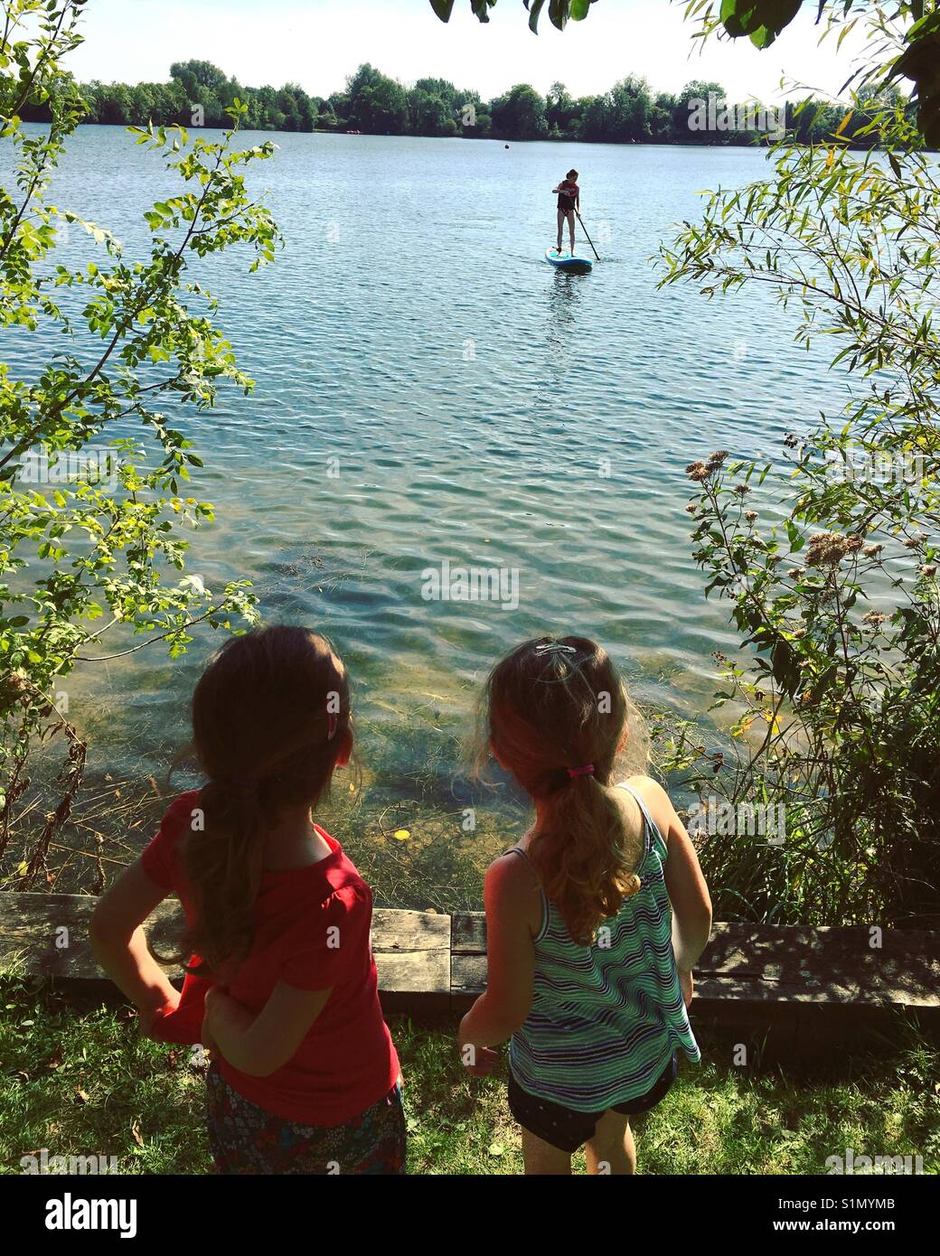 Little girls watching paddle boarder on lake Stock Photo