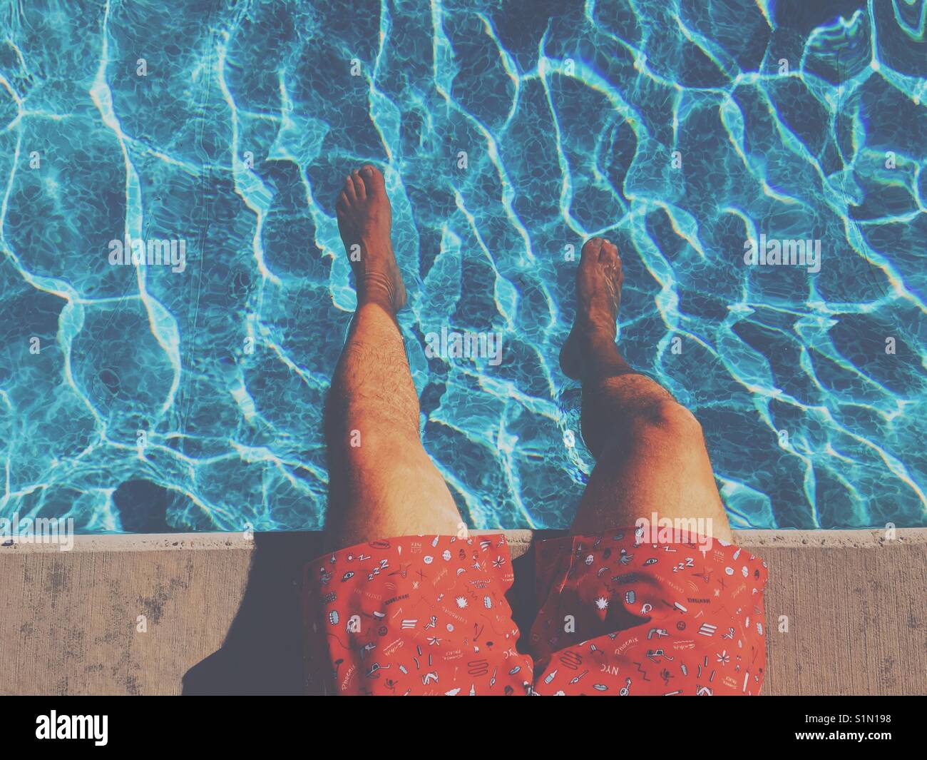 Areal view of the legs of a man sitting at the edge of a swimming pool on a hot day. - Smartphone Captured Stock Image