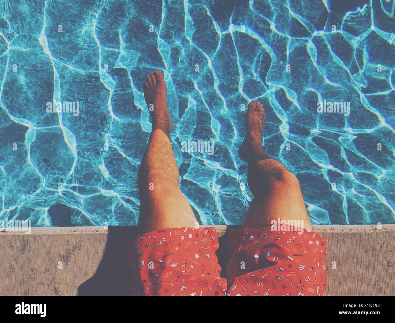 Areal view of the legs of a man sitting at the edge of a swimming pool on a hot day. Stock Photo