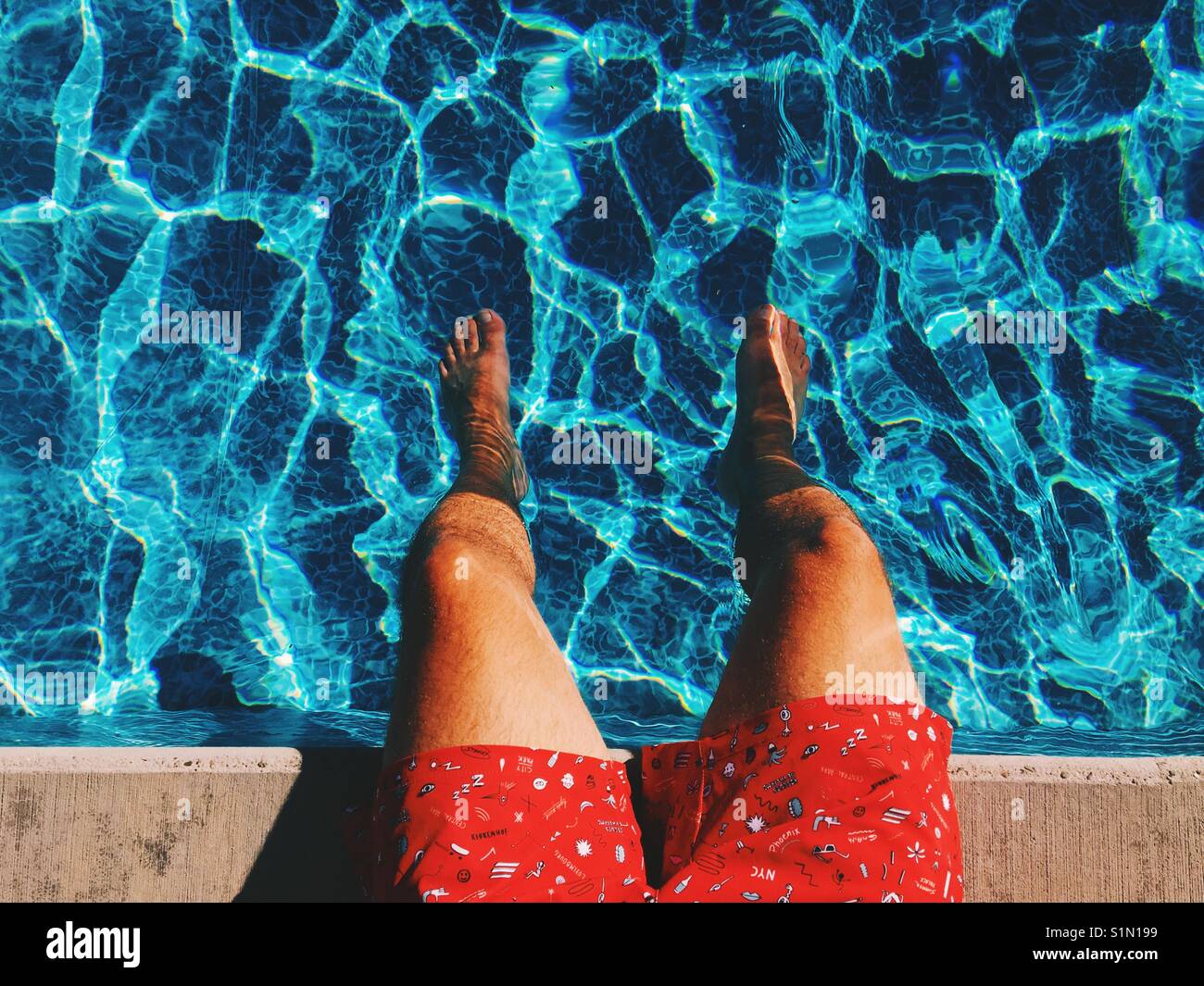 Areal view of man's legs dangling into the blue waters of a swimming pool on a sunny hot day. Stock Photo