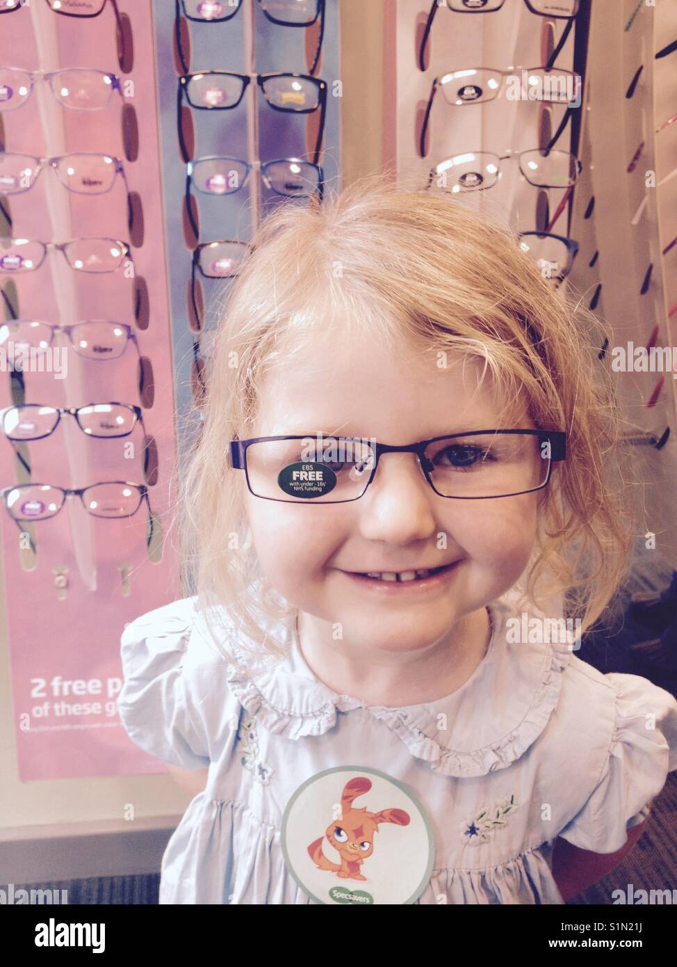 Three year old girl tries new glasses / spectacles on at an optician store. Stock Photo