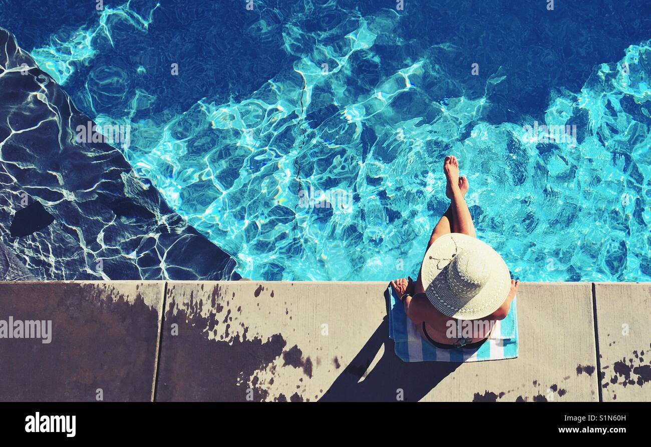 Woman wearing a large sun hat sitting at the edge of a swimming pool on a sunny summer day. Areal view. Stock Photo
